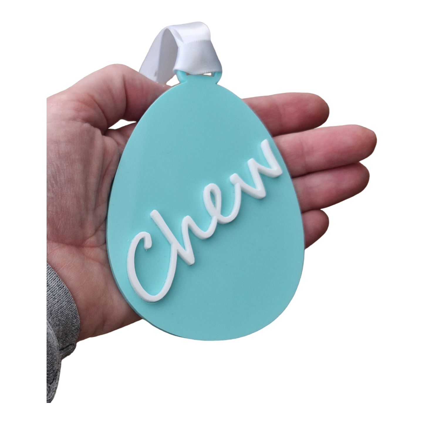 A hand holding an egg shape in the colour Green Spearmint with the name Chew going from the bottom to the top on an angle in white. With a white ribbon for hanging