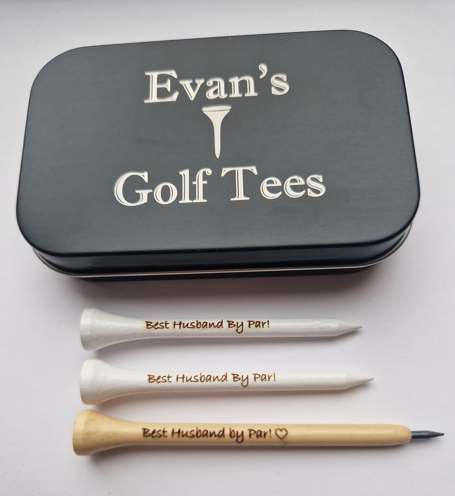 A black tin with the text "Evan's Golf Tees" and the image of a golf tee laser engraved into it. 2 white 70mm golf tees with the text "Best Husband By Par!" laser engraved into them and a wooden golf pencil shaped like a golf tee with the text "Best Husband by Par!" and the outline of a heart laser engraved into it.
