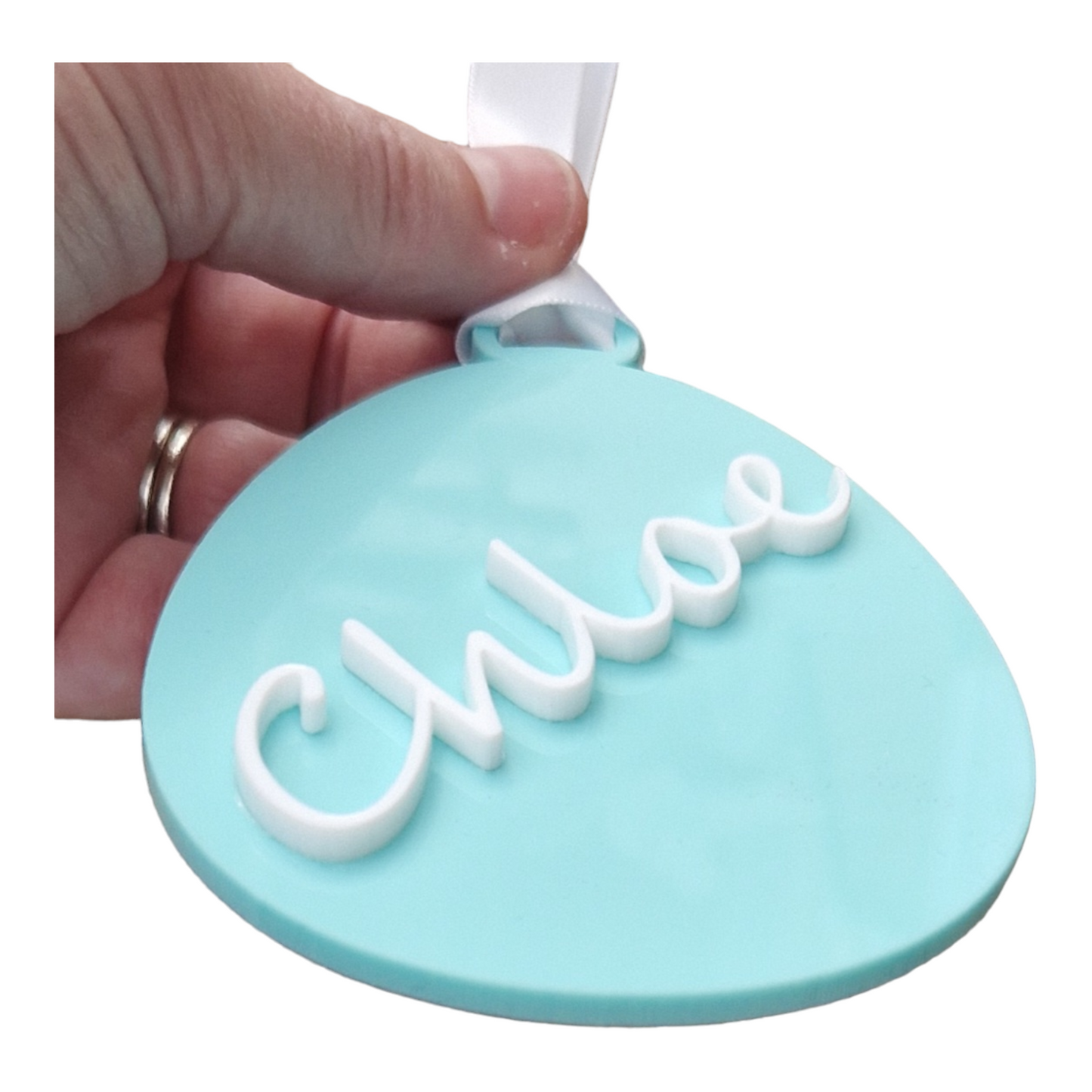 A hand holding an egg shape in the colour Green Spearmint with the name Chloe in white going from the bottom to the top at at angle, with a ribbon for hanging. The Easter decoration is held at an angle to show that the name is 3D.