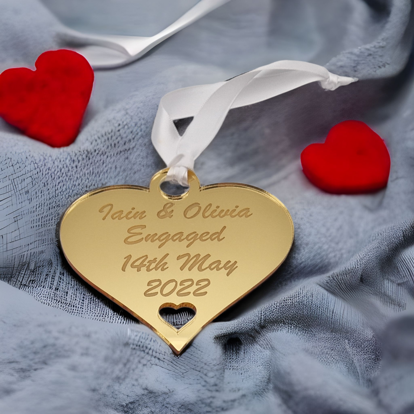 Gold heart laser engraved with the text "Iain & Olivia Engaged 14th May 2022" with a heart cut out at the bottom and a circle at the top with white ribbon for hanging. Placed on grey fabric with 2 red heart either side of it.