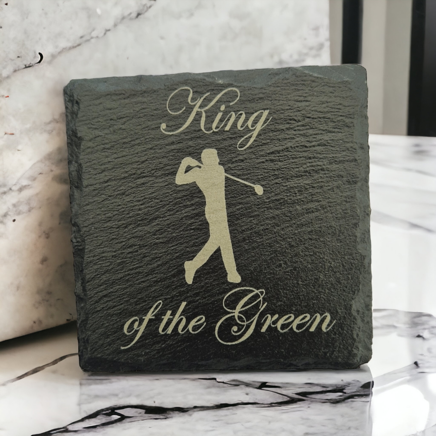 Slate coaster with the text "King of the Green" and a male golfer laser engraved on to it. The coaster is propped up on a marble worktop