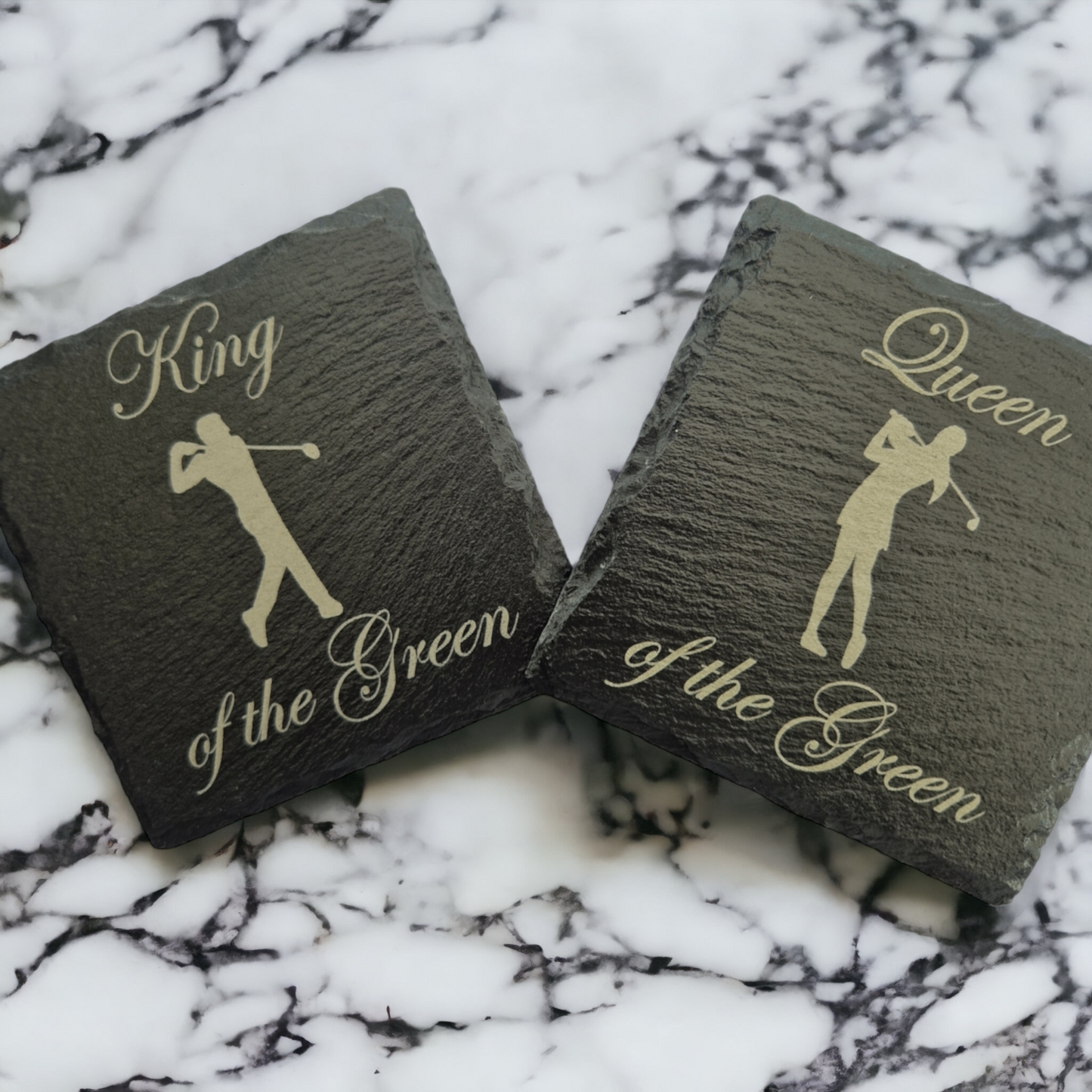 2 slate coasters - the lefthand coaster has the text "King of the Green" and the image of a male golfer. The righthand coaster has the text "Queen of the Green" and the image of a female golfer. The coasters are placed on a marble worktop