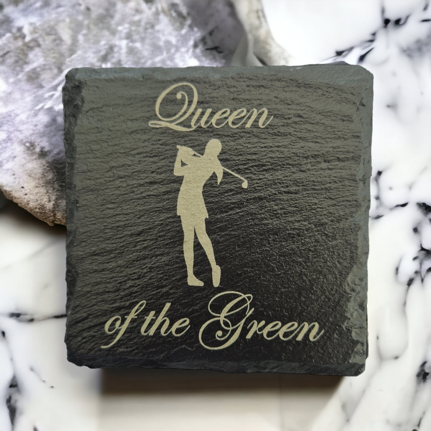 Slate coaster with the text "Queen of the Green" and the image of a female golfer laser engraved on to it. The coaster is laid on a marble worktop.