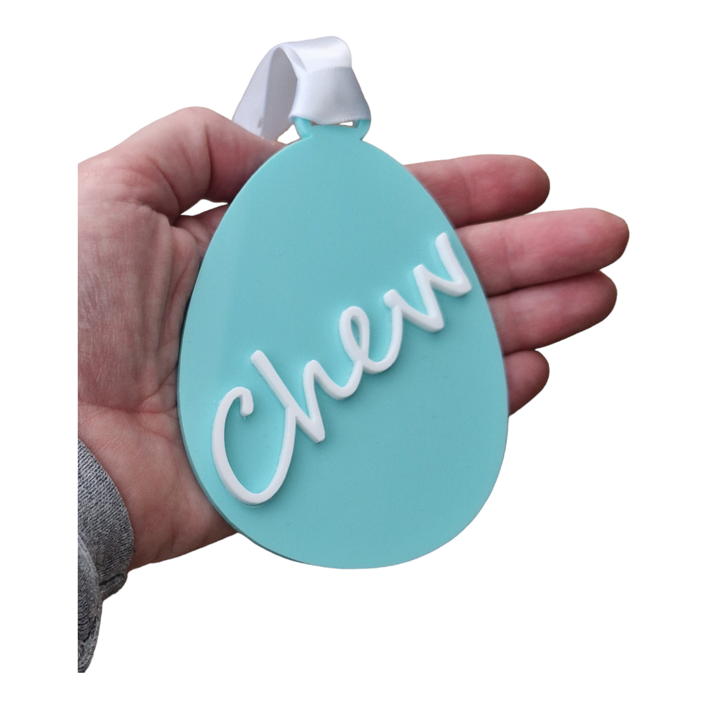 A  hand holding an egg shape in the colour Green Spearmint with the name Chew going from the bottom to the top on an angle in white. With a white ribbon for hanging