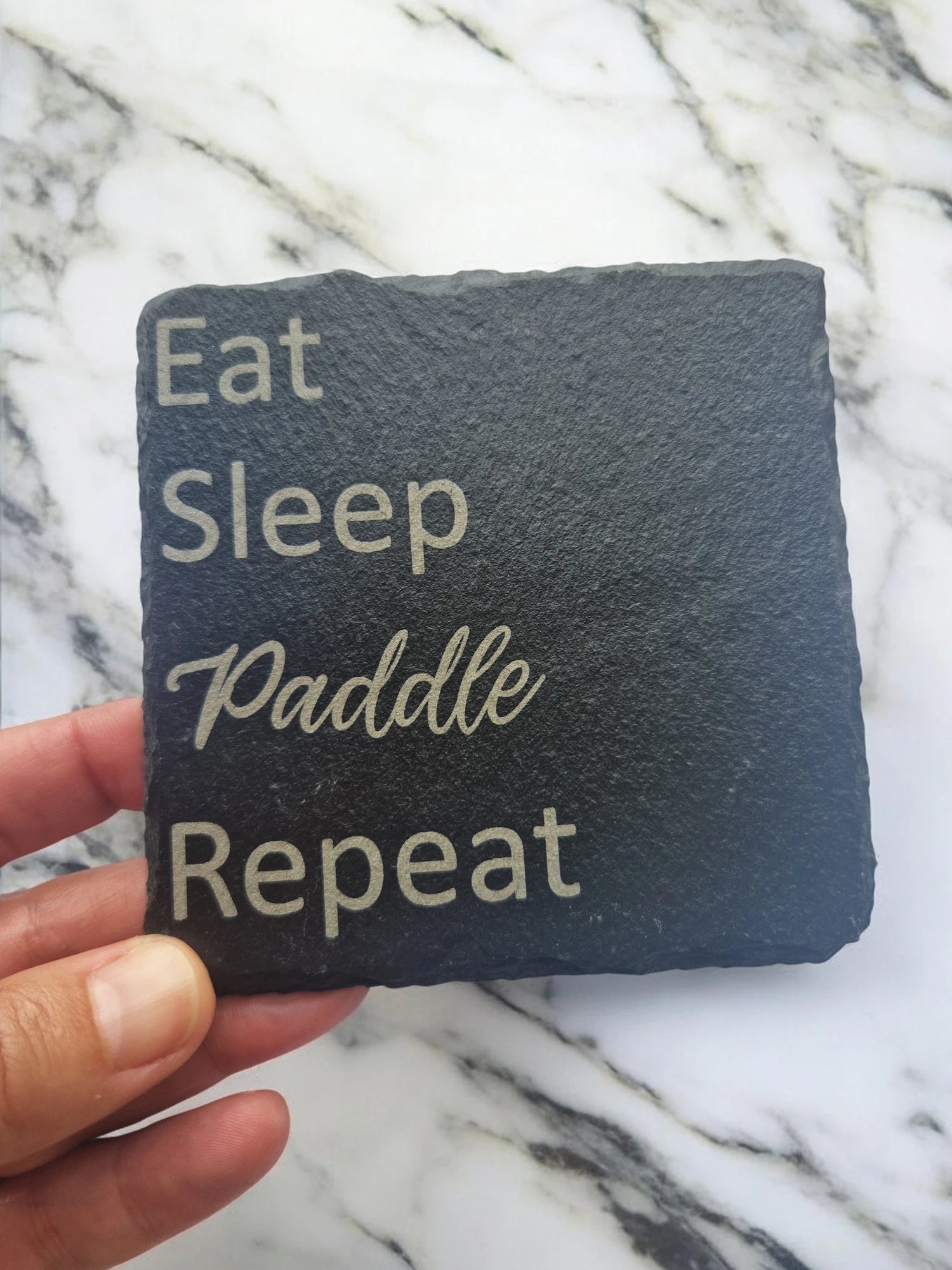 A hand holding a square coaster featuring the text Eat Sleep Paddle Repeat. Each word is on its own line, Eat Sleep Repeat are in a standard font, Paddle is in a cursive font.