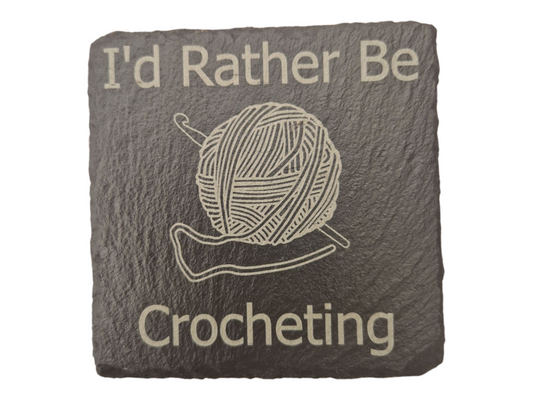 Square slate coaster with the text "I'd Rather Be" at the top of the coaster and "Crocheting" at the bottom of the coaster. In the centre of the coaster is the image of a ball of wool with a crochet hook going diagonally through it.