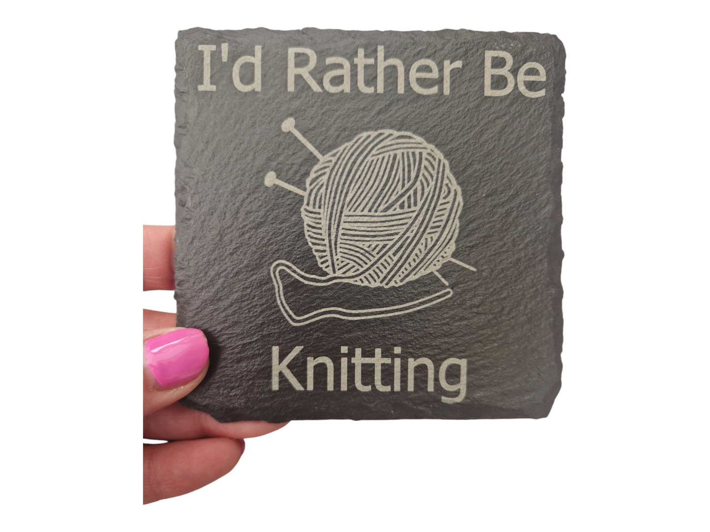 A hand holding a square slate coaster with the text "I'd Rather Be" at the top of the coaster and "Knitting" at the bottom of the coaster. In the centre of the coaster is the image of a ball of wool with two knitting needles going diagonally through it.