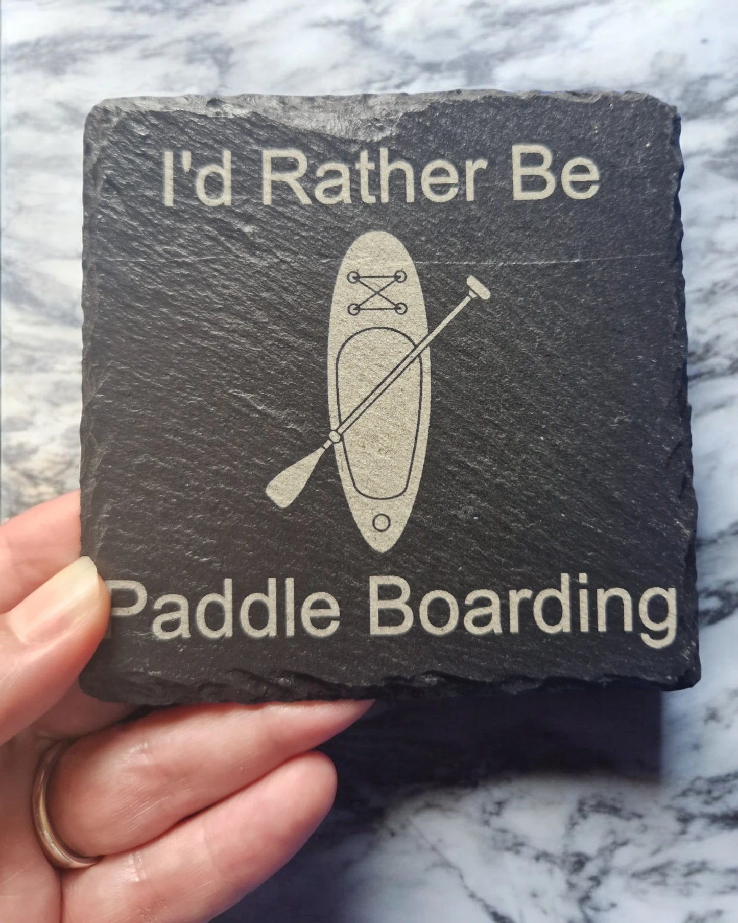 a hand holding a black slate coaster with the text "I'd Rather Be" at the top of the coaster and "Paddle Boarding" at the bottom of the coaster. In the centre is the image of a paddle board with a paddle placed diagonally across it. 