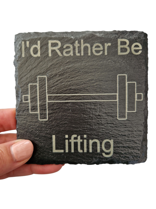 A hand holding a square slate coaster with the text "I'd Rather Be Lifting" and the image of a barbell in the centre of the coaster. 