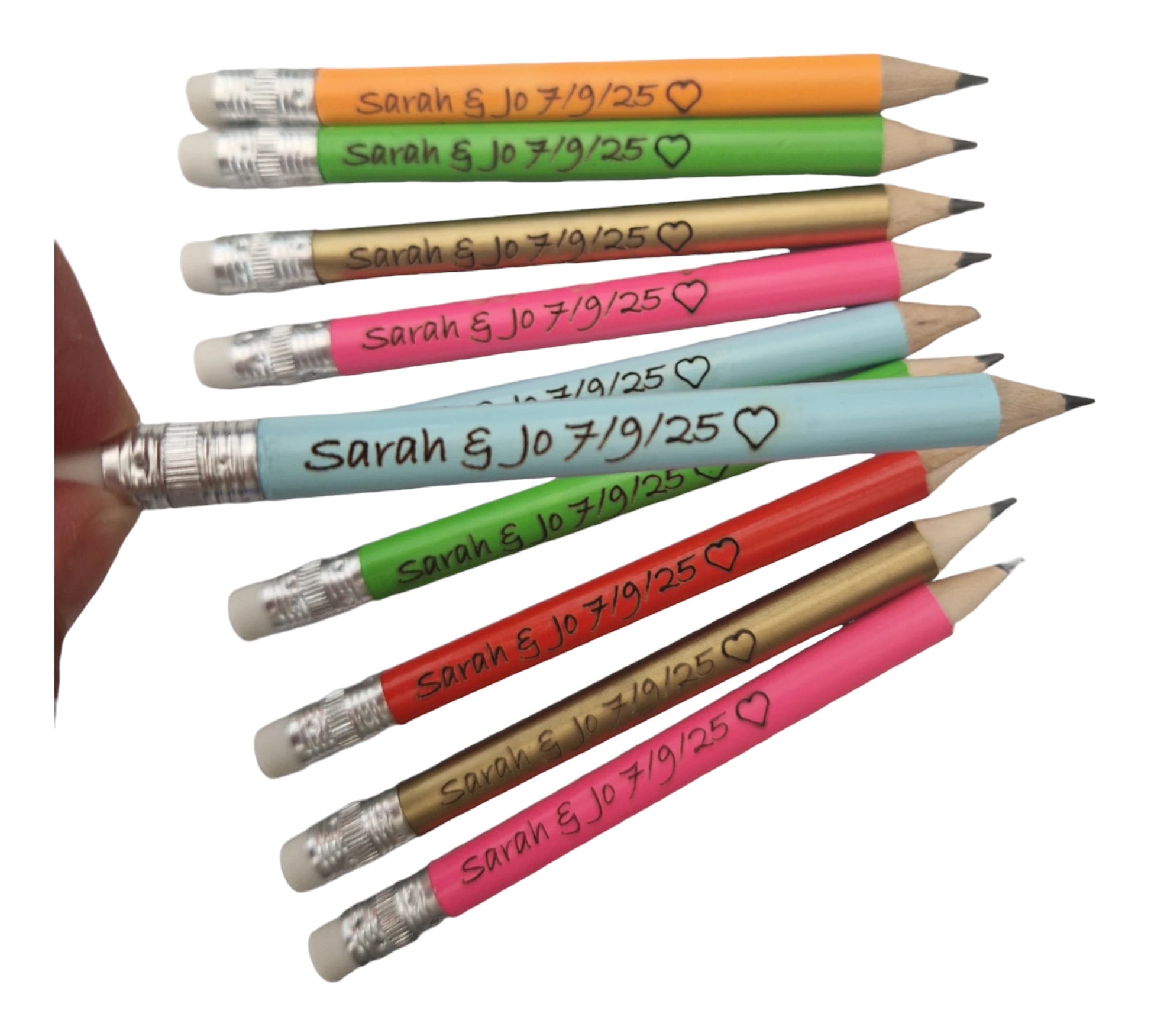 neon orange, neon green, gold, neon pink, light blue and red pencils with the text "Sarah & Jo 7/9/25" and the outline of a heart laser engraved into them. A light blue pencil is being held closer o the camera to show the engraving more clearly.