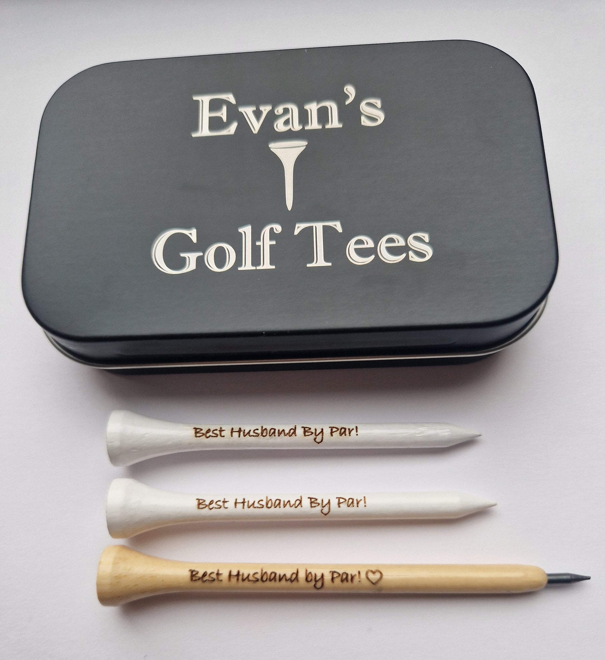 A black tin with the text "Evan's Golf Tees" and the image of a golf tee laser engraved into it. 2  white 70mm golf tees with the text "Best Husband By Par!" laser engraved into them and a wooden golf pencil shaped like a golf tee with the text "Best Husband by Par!" and the outline of a heart laser engraved into it. 