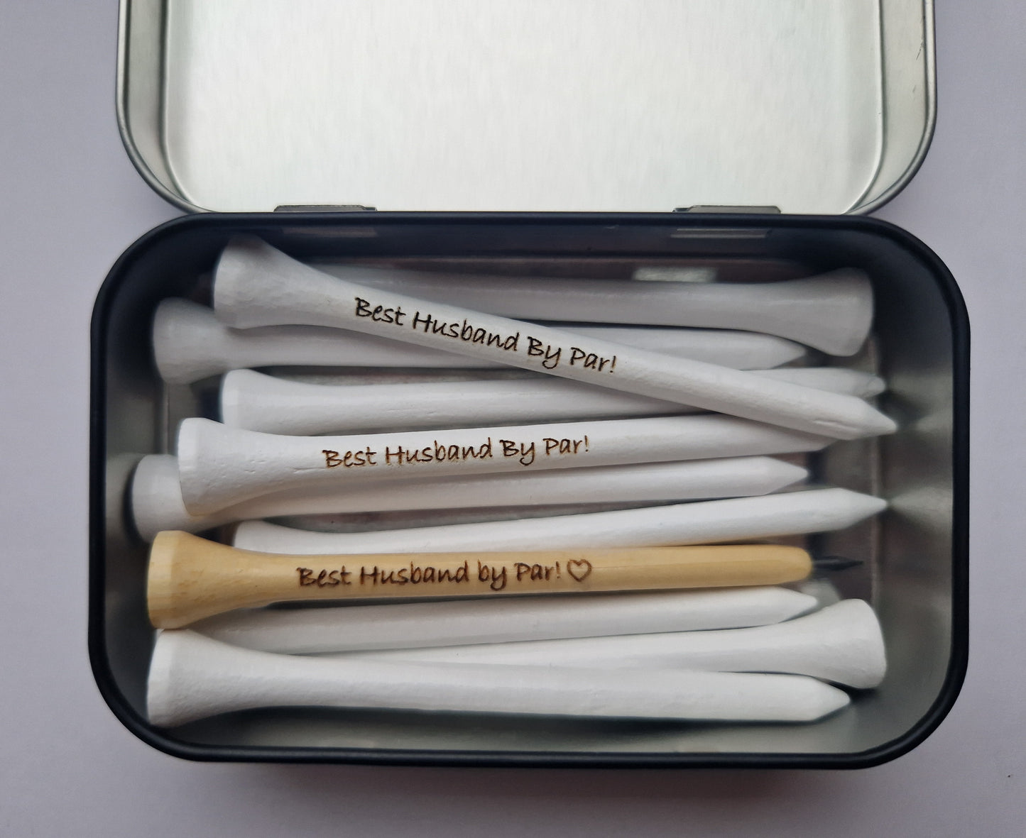 An open tin with 10 white 83mm golf tees laser engraved with the text "Best Husband By Par!" A wooden pencil shaped like a golf tee with pencil lead at the tip with the text "Best Husband by Par!" and the outline of a heart laser engraved into it.