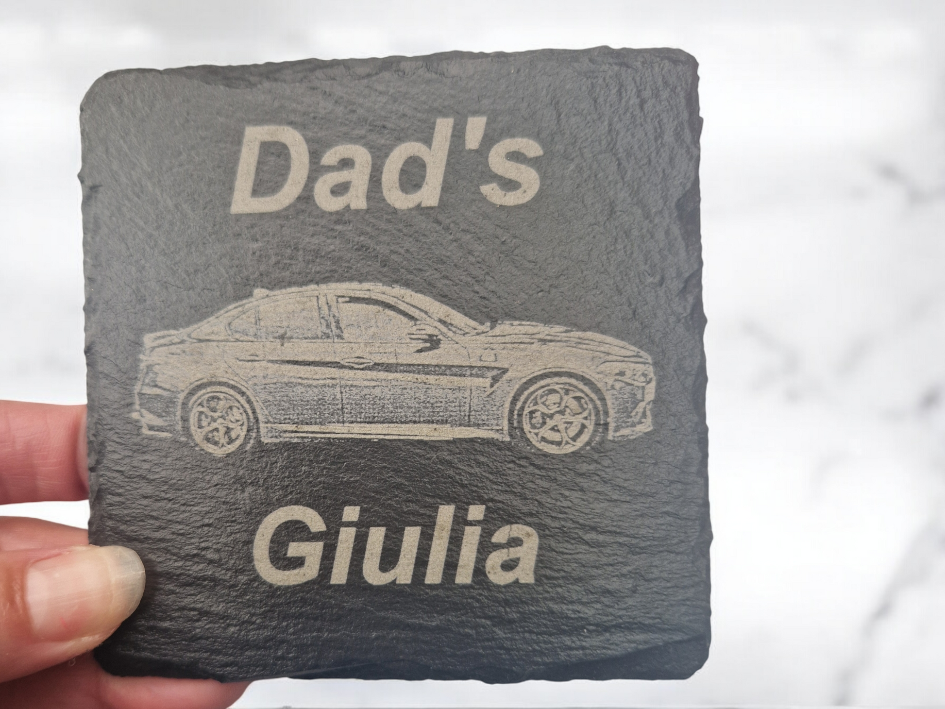 A hand holding a square slate coaster featuring the text "Dad's" at the top of the coaster and "Giulia" at the bottom of the coaster. In the centre of the coaster is a laser engraved image of a car.