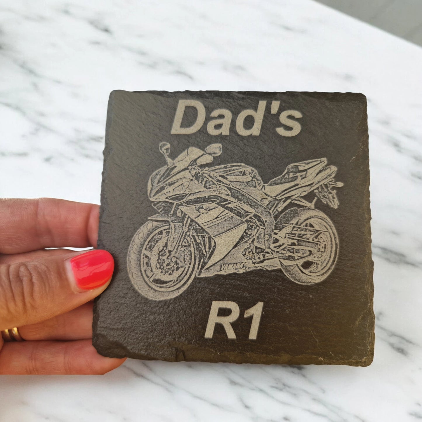 Square slate coaster with a motorbike laser engraved into the centre of the coaster. At the top of the coaster is the text "Dad's" at the bottom of the coaster is the text "R1". The coaster is being held against a marble worktop.