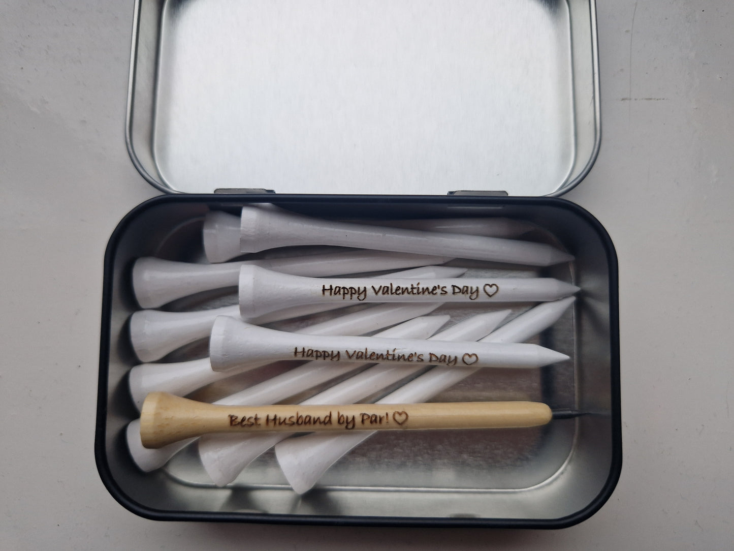 An open tin with 10 white 70mm golf tees with the text "Happy Valentine's Day" and the image of a heart laser engraved into them. A wooden golf tee pencil shaped like a golf tee with pencil lead at the tip with the text "Best Husband by Par!" and the outline of a heart laser engraved into it. 