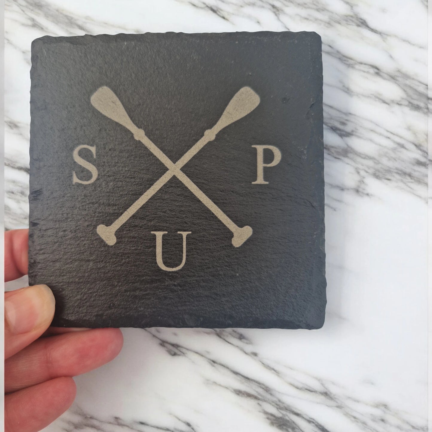 A hand holding a slate coaster laser engraved with 2 paddles crossed over with the letter "S" to the left of the paddles, the letter "U" under the paddles and the letter "P" to the right of the paddles