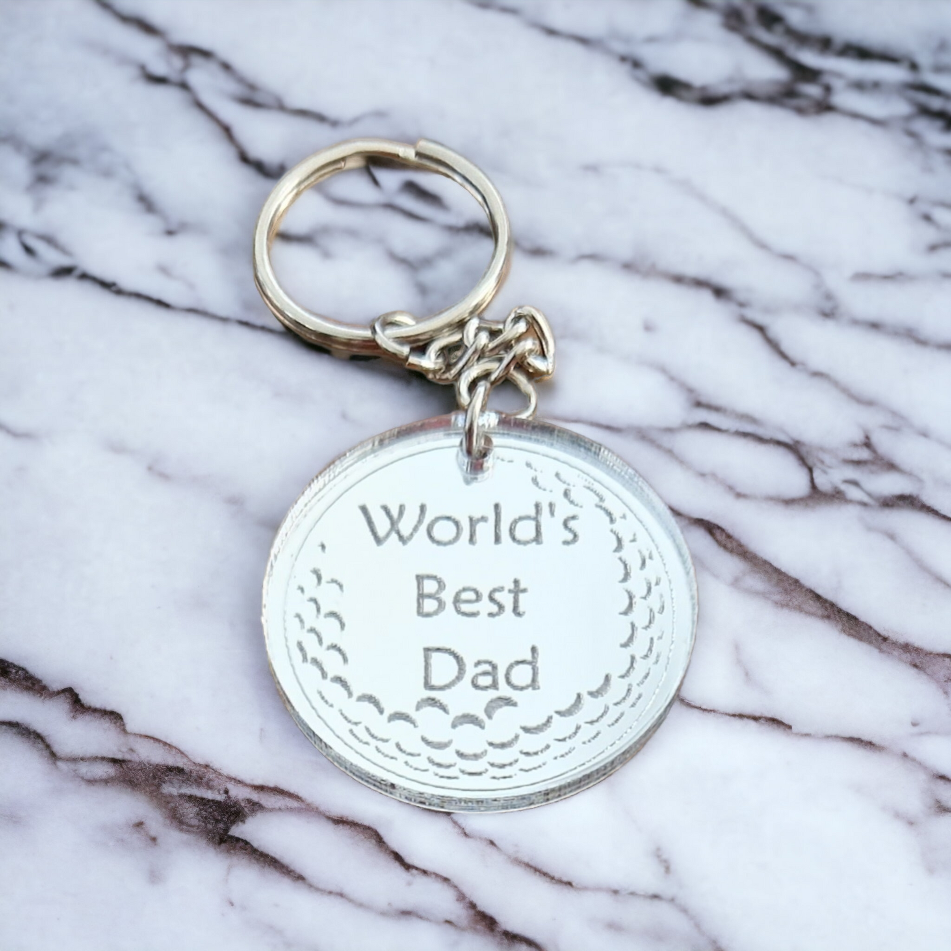 Mirror silver circle with the partial image of a golf ball and the text "World's Best Dad" laser engraved into it. The circle is attached to a metal keychain. The keyring is on a marble work top