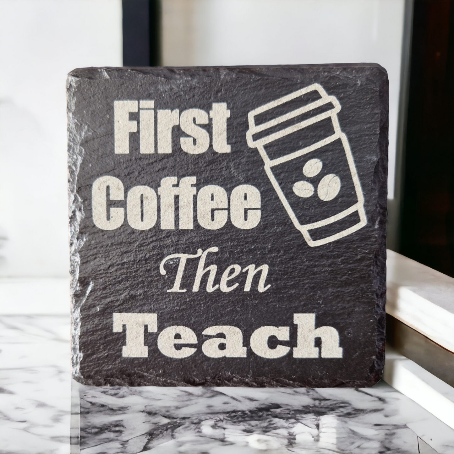 Slate coaster with the text "First Coffee then Teach" and the image of a take away coffee cup with coffee beans on it. Propped up on a marble work top