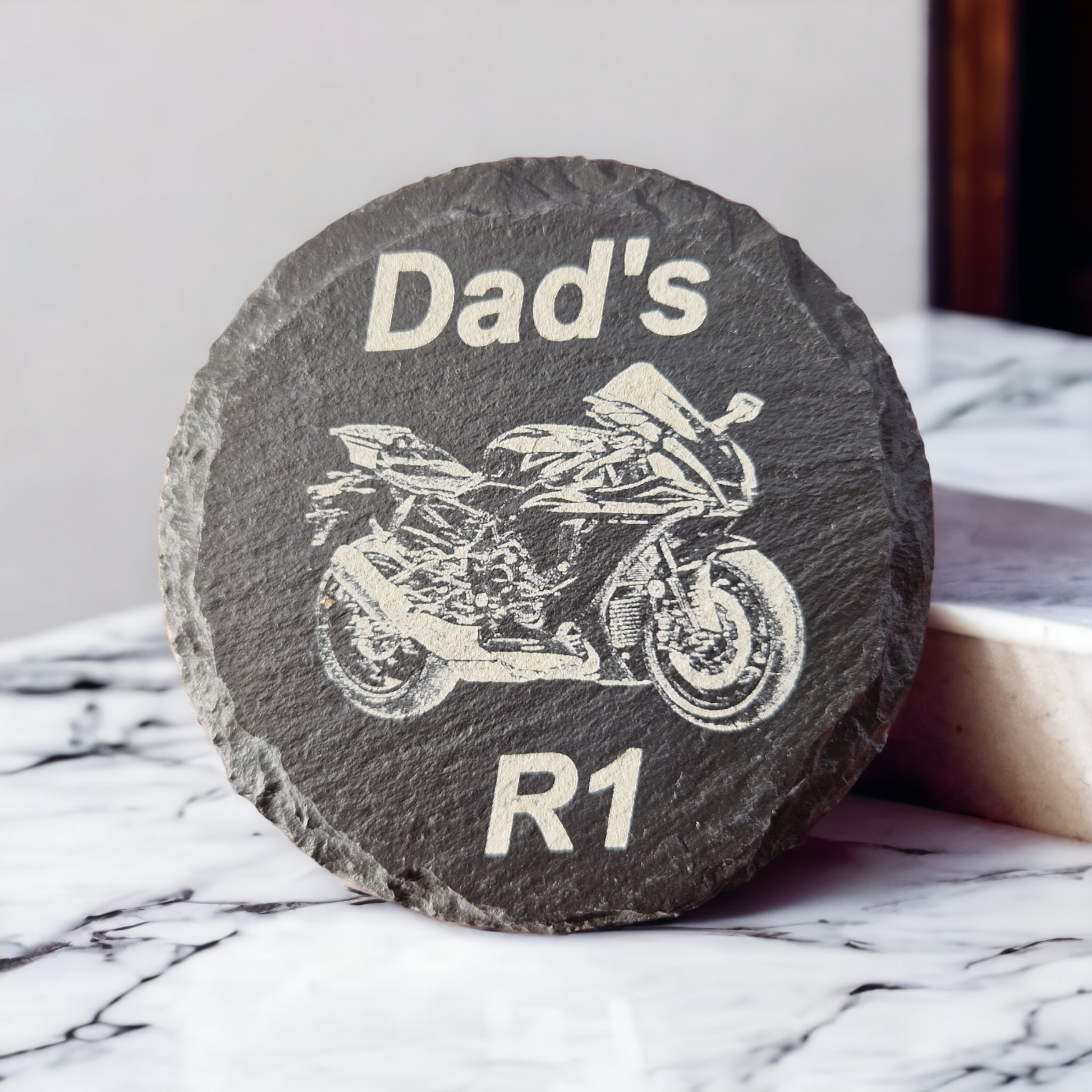 Round slate coaster with the text "Dad's" at the top  of the coaster and "R1" at the bottom of the coaster. With the image of a Yamaha R1 laser engraved into the centre of the coaster. The coaster is propped up on a marble work top.