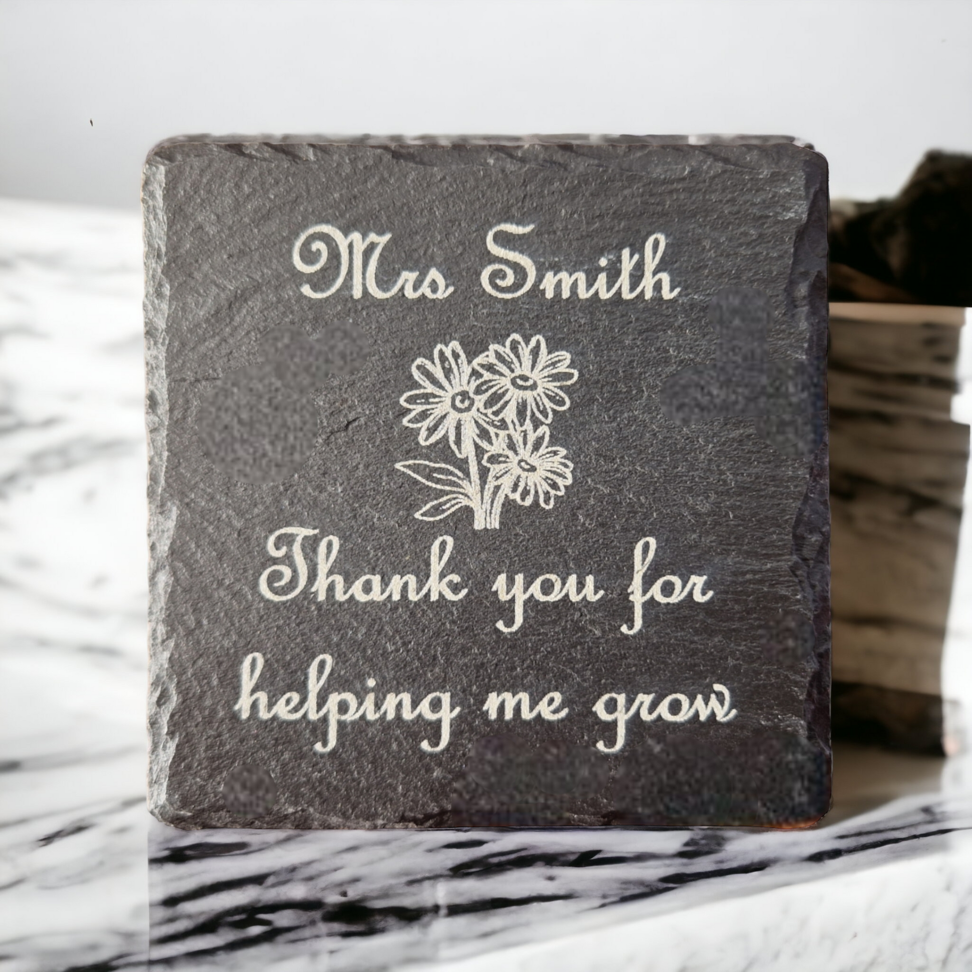 Slate coaster with the text "Mrs Smith Thank you for helping me grow" and the image of some flowers in the centre of the coaster. Propped up on a marble work top.