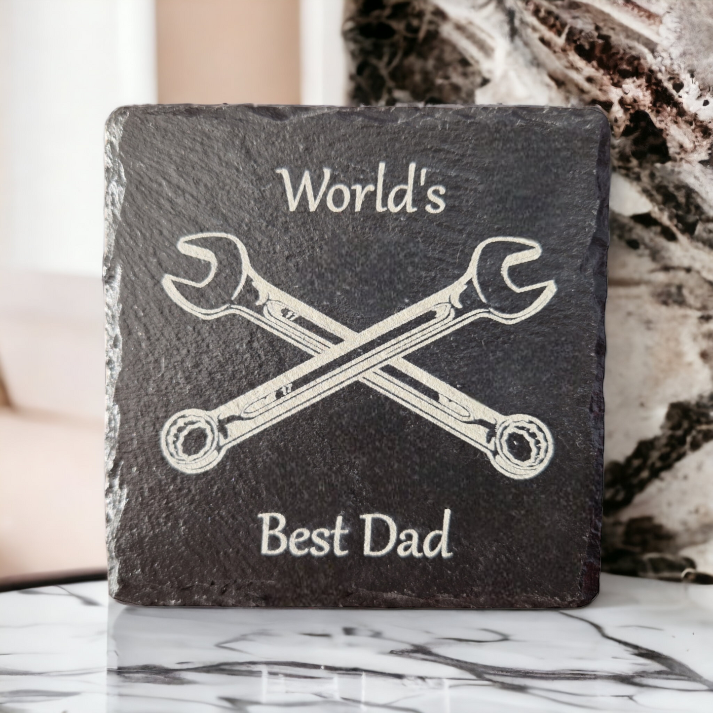 Square slate coaster with the text "World's" at the top of the coaster and "Best Dad" at the bottom of the coaster. In the centre of the coaster are two spanners crossed over to form an "X" shape. The coaster is stood up on a marble work top