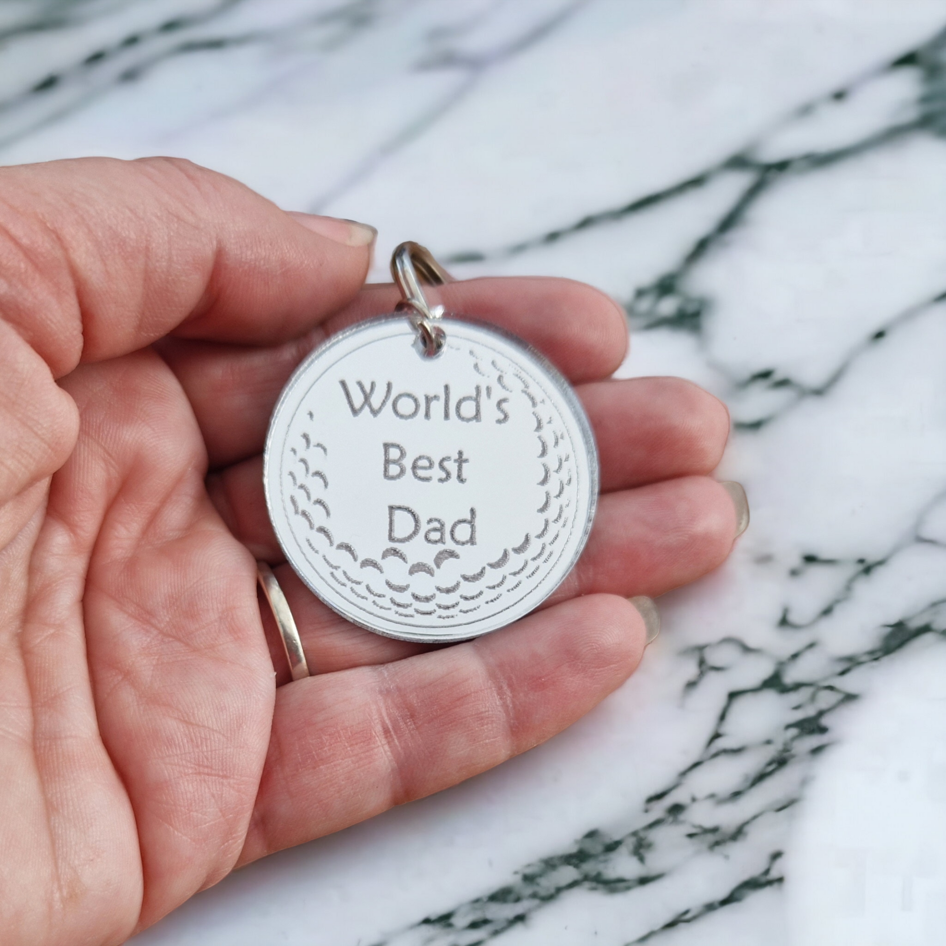 Mirror silver circle with the partial image of a golf ball and the text "World's Best Dad" laser engraved into it. The circle is attached to a metal keychain. The keyring is being held in a hand over a marble work top