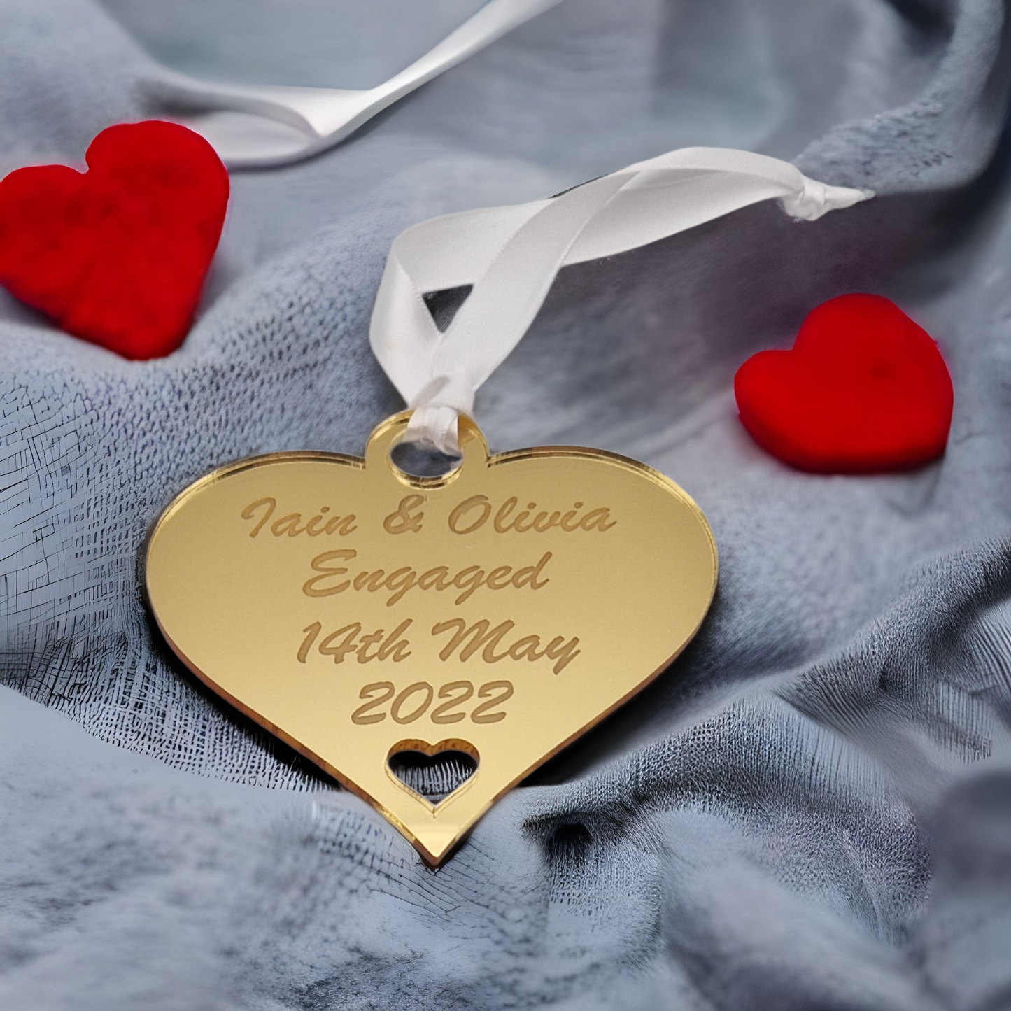 Gold heart laser engraved with the text "Iain & Olivia Engaged 14th May 2022" with a heart cut out at the bottom and a circle at the top with white ribbon for hanging. Placed on grey fabric with 2 red heart either side of it. 