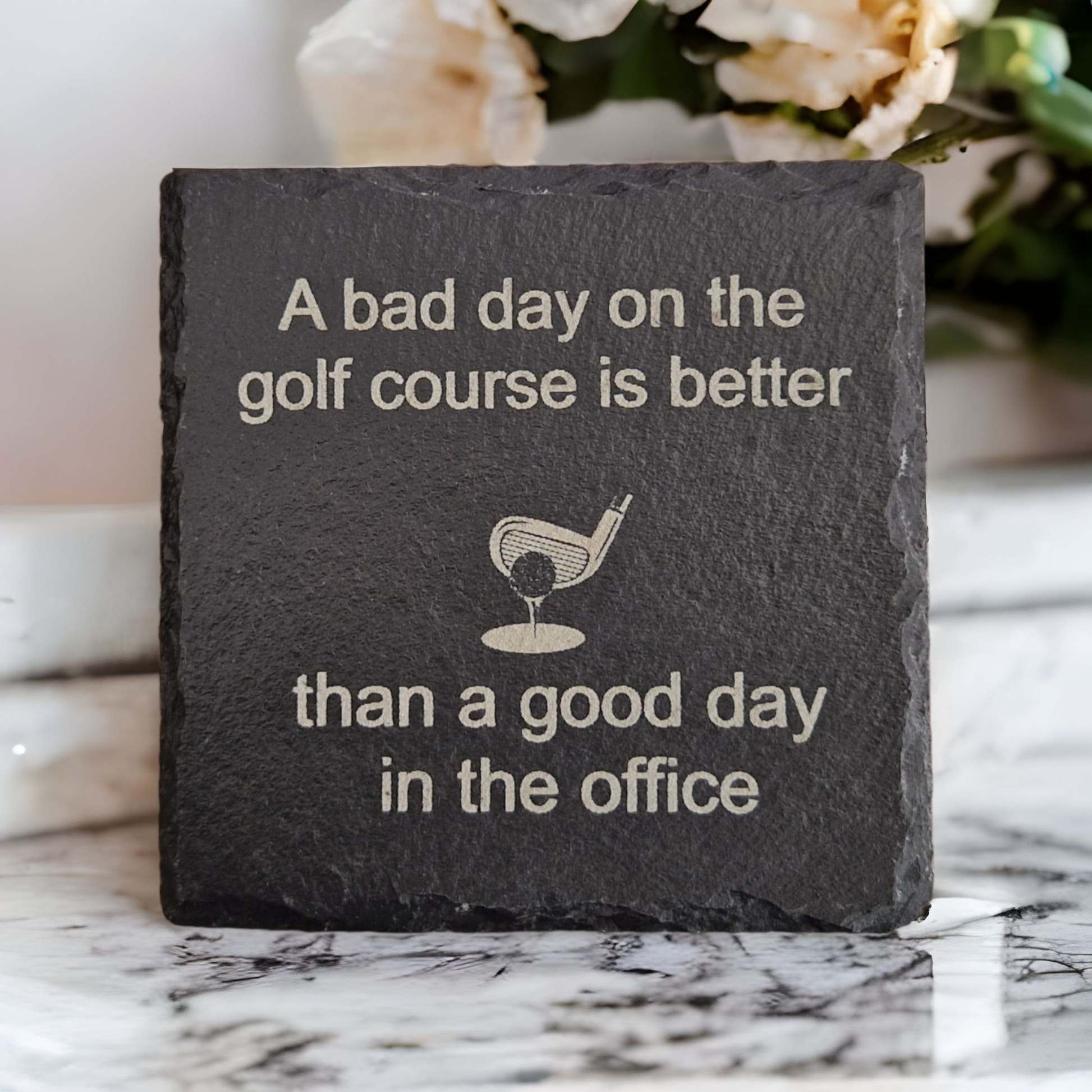 Laser engraved coaster featuring the text "A bad day on the golf course is better" at the top of the coaster, "than a good day in the office" at the bottom of the coaster. In the centre of the coaster is a golf ball on a tee about to be hit by a club. The coaster is sat on a marble worktop with flowers in the background