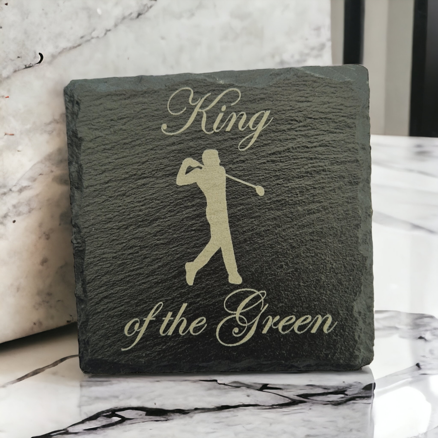 Slate coaster with the text "King of the Green" and a male golfer laser engraved  on to it. The coaster is propped up on a marble worktop