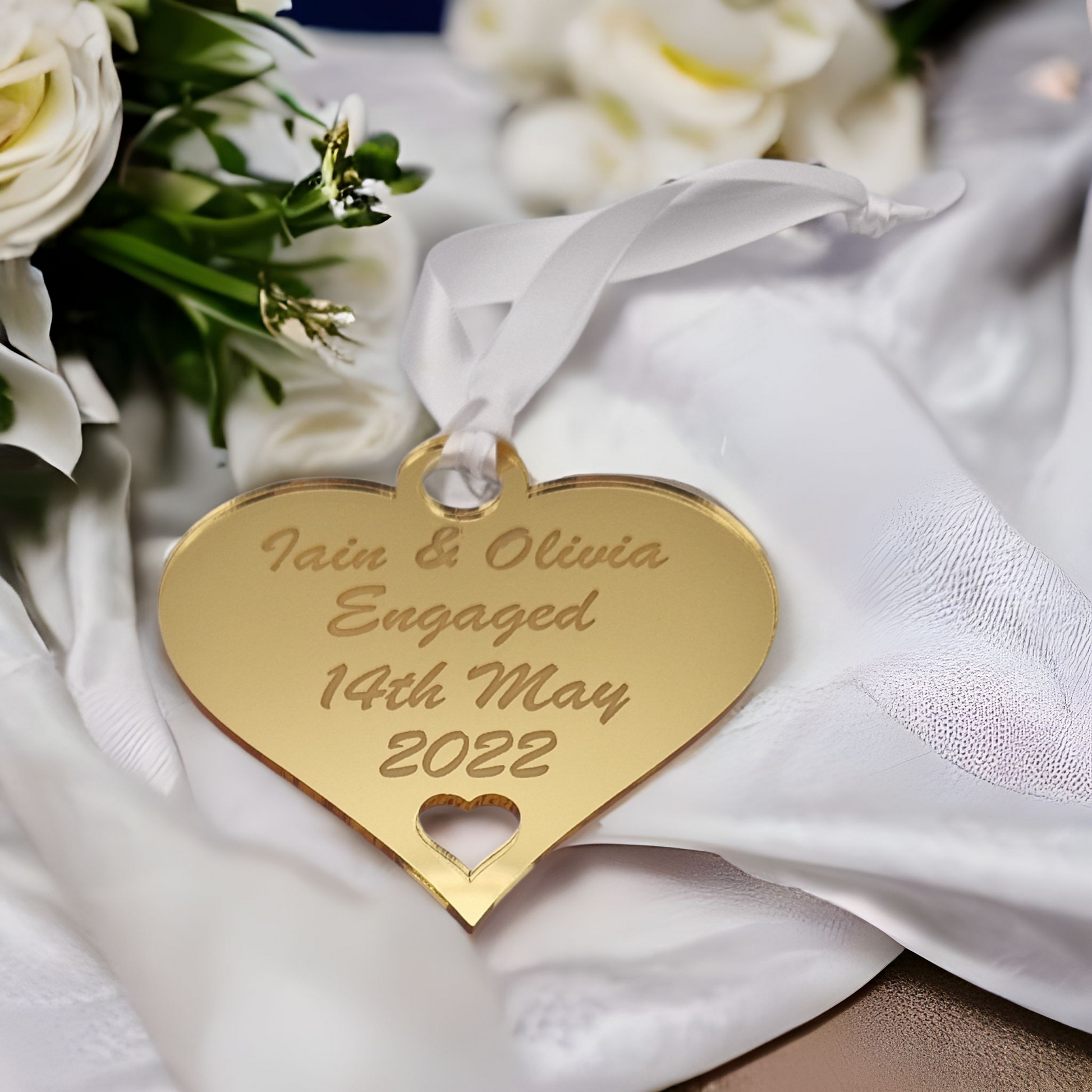 Gold heart laser engraved with the text "Iain & Olivia Engaged 14th May 2022" with a heart cut out at the bottom and a circle at the top with white ribbon for hanging. Laid on white fabric with flowers in the background.