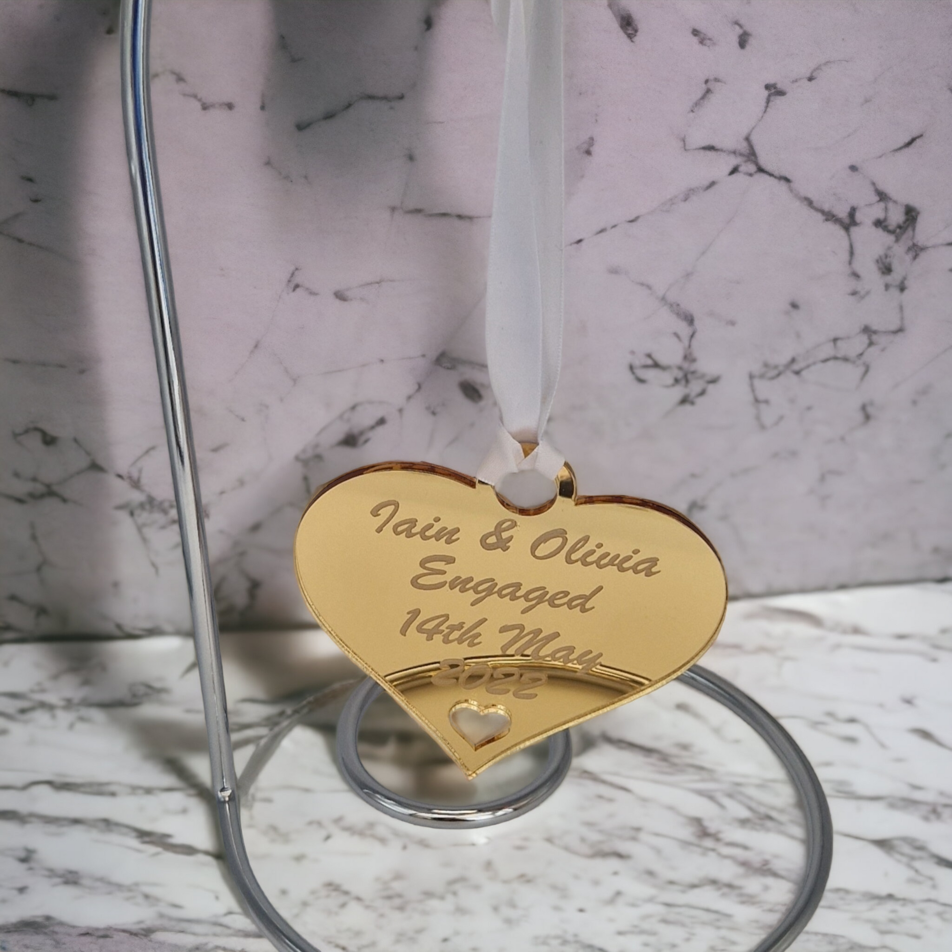 Gold heart laser engraved with the text "Iain & Olivia Engaged 14th May 2022" with a heart cut out at the bottom and a circle at the top with white ribbon for hanging. The heart is hung on a metal hook stand that is placed on a marble worktop.