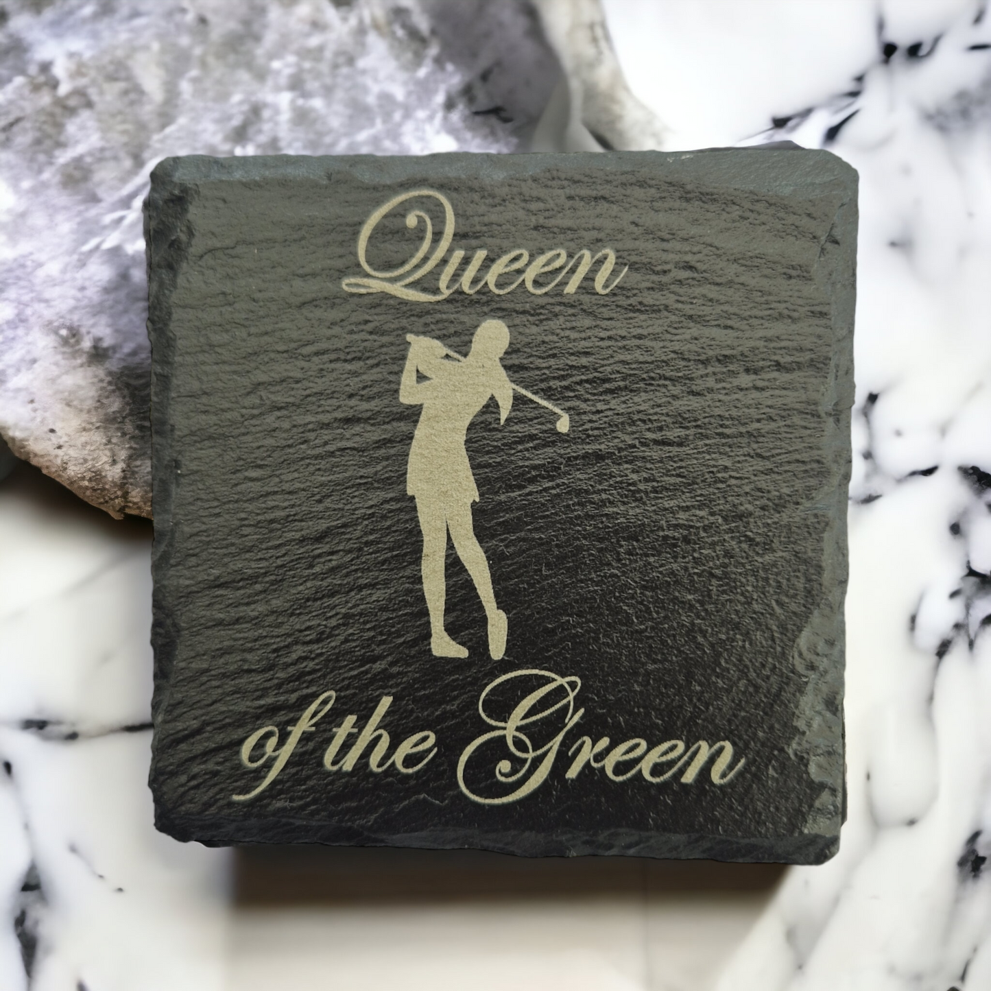 Slate coaster with the text "Queen of the Green" and the image of a female golfer laser engraved on to it. The coaster is laid on a marble worktop. 