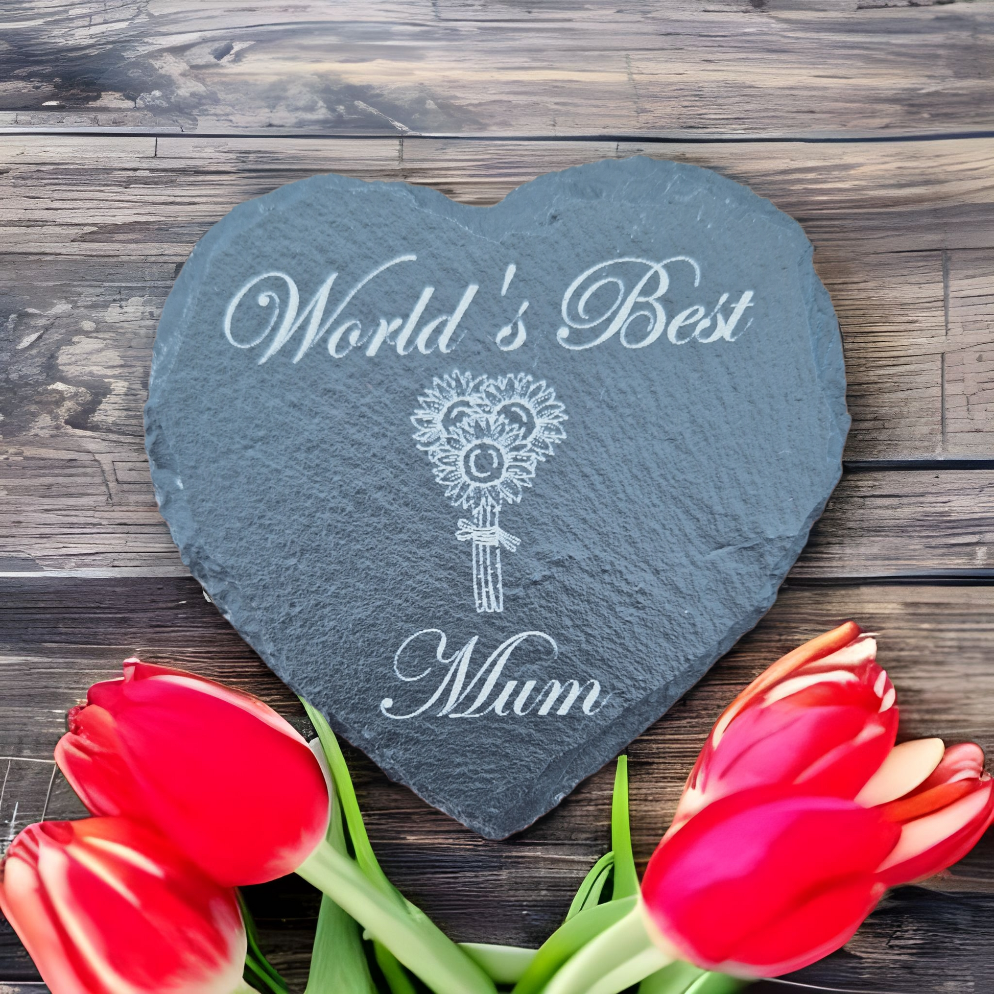 Slate heart coaster with the text "World's Best" at the top of the coaster and "Mum" and the bottom. In the centre there are 3 sunflowers arranged into a bouquet. Placed on a wooden bench with tulips at the base of the coaster