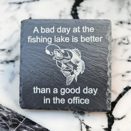 Slate coaster featuring the text "A bad day at the fishing lake is better" laser engraved at the top of the coaster, "than a good day in the office" laser engraved at the bottom of the coaster. In the centre of the coaster is a carp almost getting caught by a fishing line. The coaster is placed on a marble worktop.