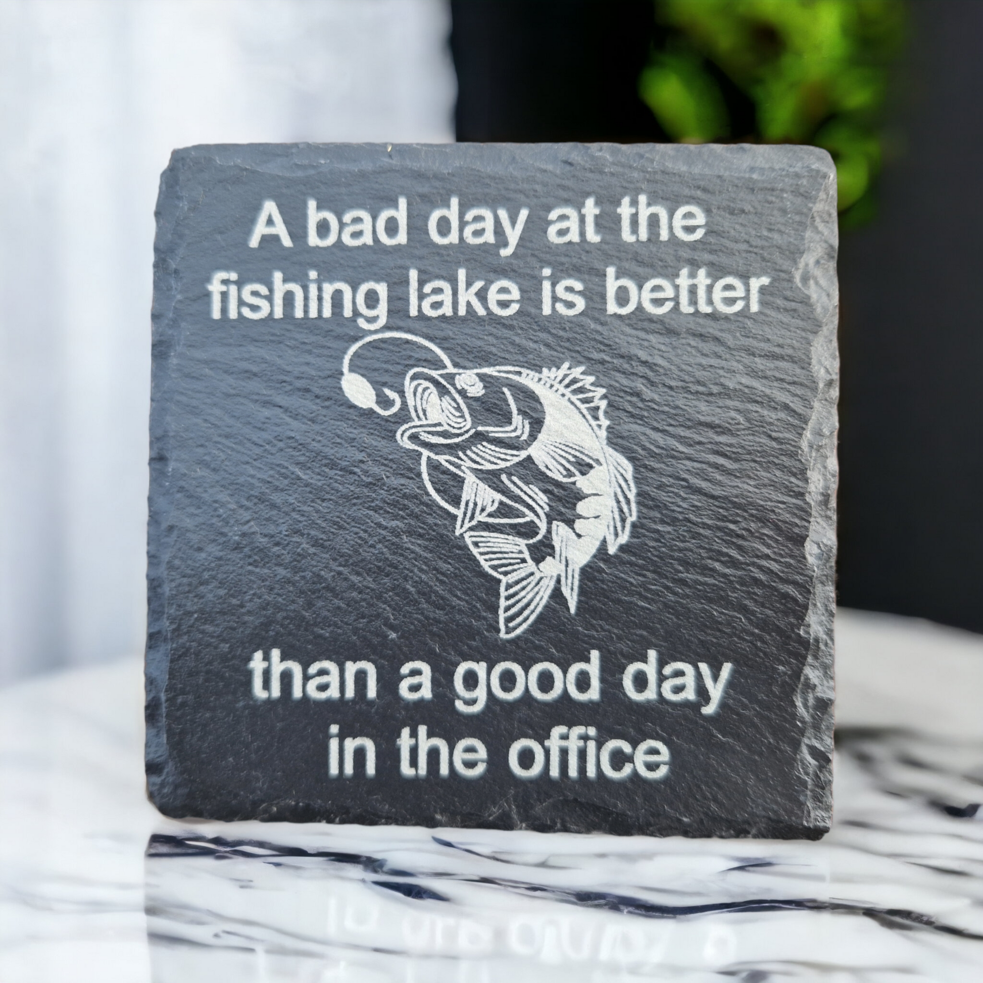 Slate coaster featuring the text "A bad day at the fishing lake is better" laser engraved at the top of the coaster, "than a good day in the office" laser engraved at the bottom of the coaster. In the centre of the coaster is a carp almost getting caught by a fishing line. The coaster is stood on a marble worktop.