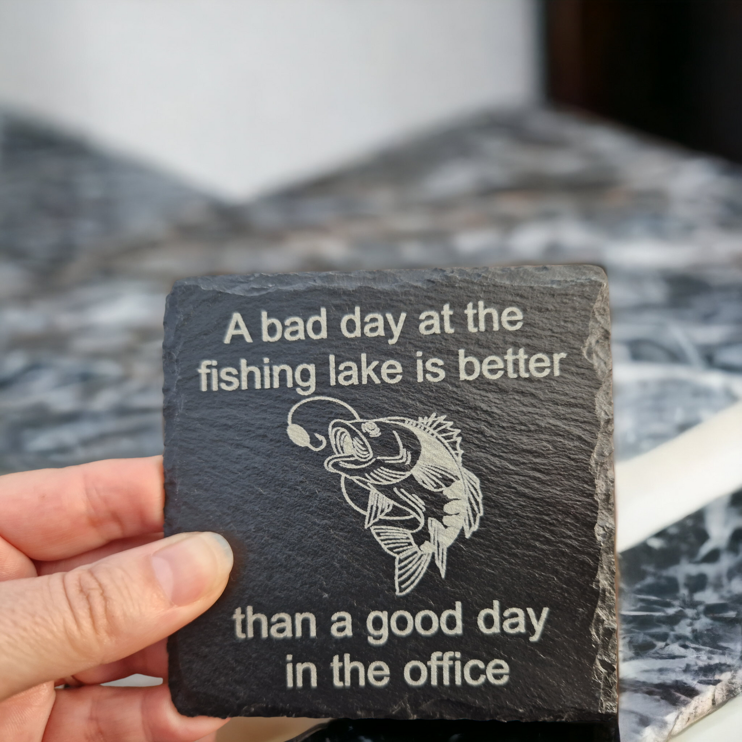 Slate coaster featuring the text "A bad day at the fishing lake is better" laser engraved at the top of the coaster, "than a good day in the office" laser engraved at the bottom of the coaster. In the centre of the coaster is a carp almost getting caught by a fishing line. The coaster is being held over a marble worktop.