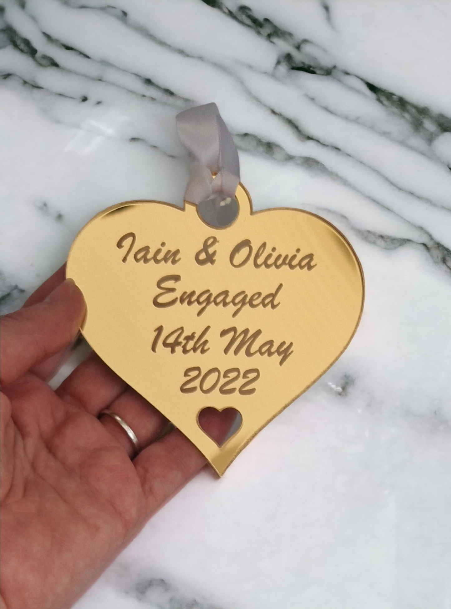 Gold heart laser engraved with the text "Iain & Olivia Engaged 14th May 2022" with a heart cut out at the bottom and a circle at the top with white ribbon for hanging. The heart is being held over a marble worktop