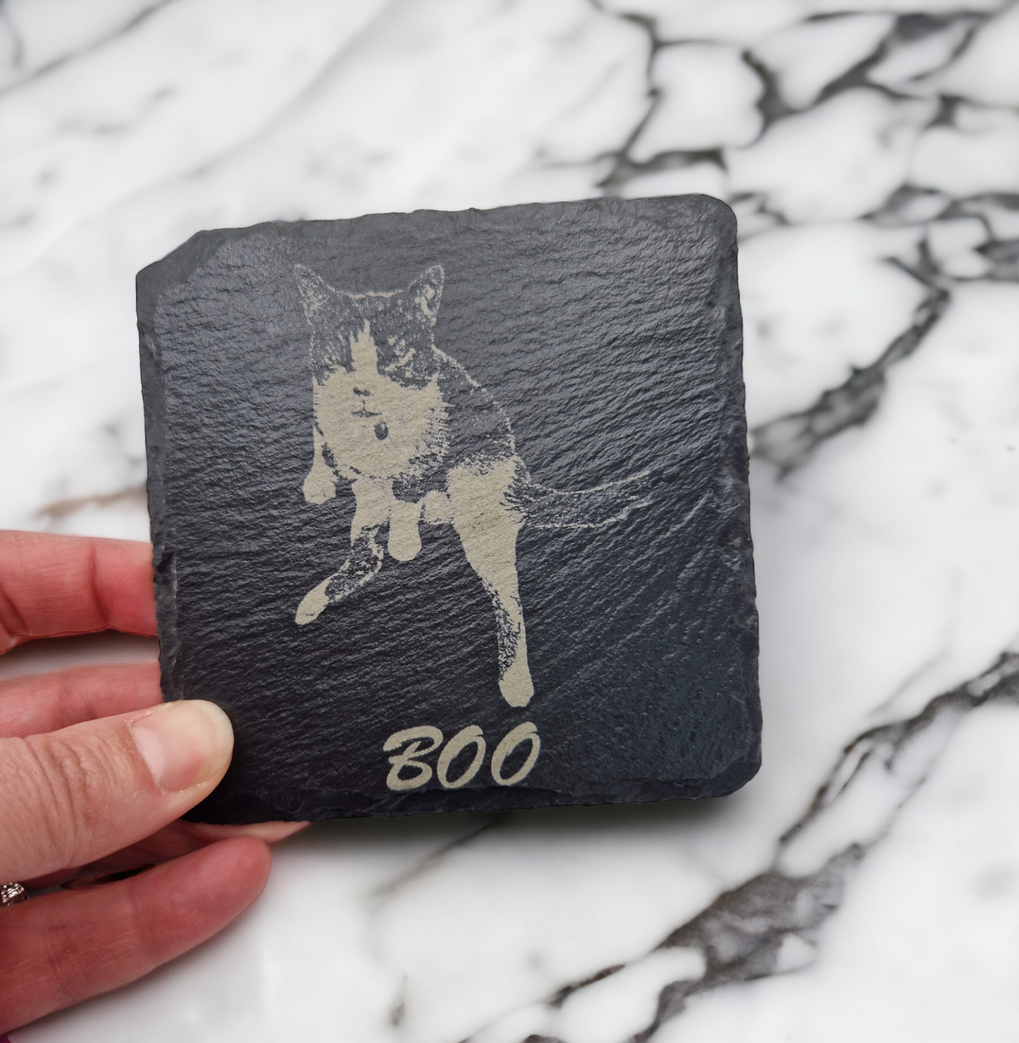 Slate coaster featuring the image of a cat laser engraved into it, with the text "Boo" written at the bottom in cursive text. The coaster is being held over a marble worktop.