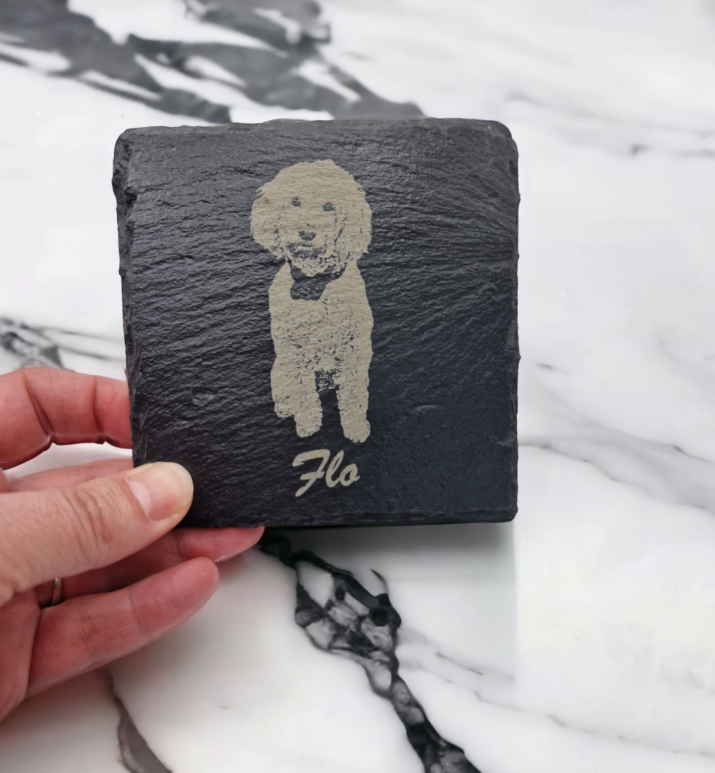 Slate coaster featuring the image of a dog laser engraved into it, with the text "Flo" written at the bottom in cursive text. The coaster is being held over a marble worktop.