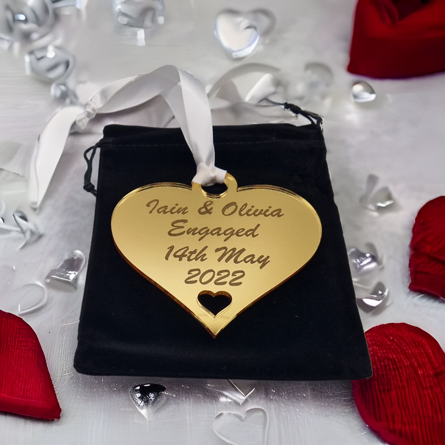 Gold heart laser engraved with the text "Iain & Olivia Engaged 14th May 2022" with a heart cut out at the bottom and a circle at the top with white ribbon for hanging. Placed on a black velvet bag, which is placed on white fabric with red, silver and clear hearts dotted around it.