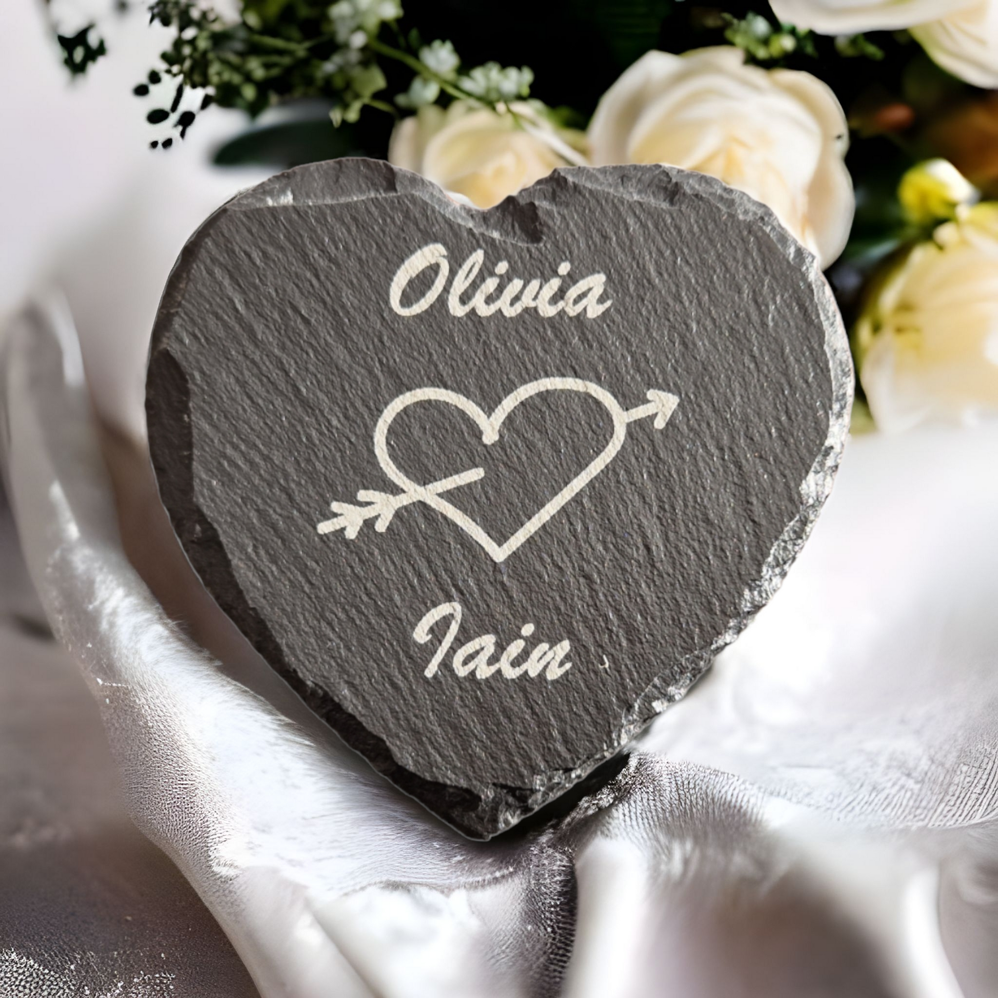 Laser engraved slate heart with "Olivia" engraved at the top and "Iain" engraved at the bottom, with the image of a heart with an arrow running through it. the coaster is on top of some white material with flowers in the background