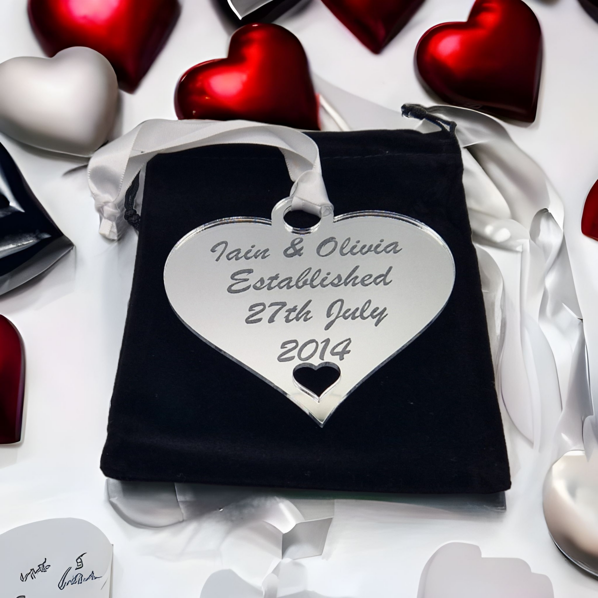 Silver acrylic heart laser engraved with the text "Iain & Olivia Established 27th July 2014" There is a heart cut out at the bottom of the hanging heart. The heart has a white satin ribbon for hanging and is sat on a black velvet bag.