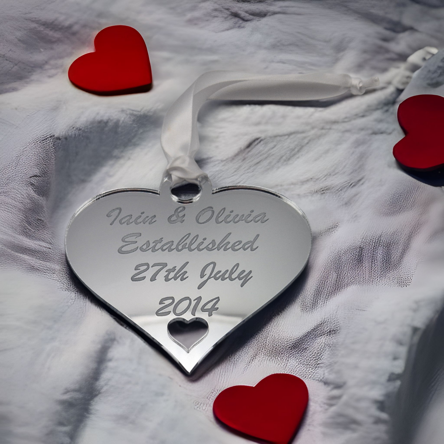 Silver acrylic heart laser engraved with the text "Iain & Olivia Established 27th July 2014" There is a heart cut out at the bottom of the hanging heart. The heart has a white satin ribbon for hanging and is sat on a white sheet with 3 red heart placed on the sheet